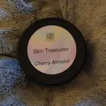 Elixian Essence Skin Treasures body cream in a blue jar with a black lid, featuring a Cherry Almond label on the lid